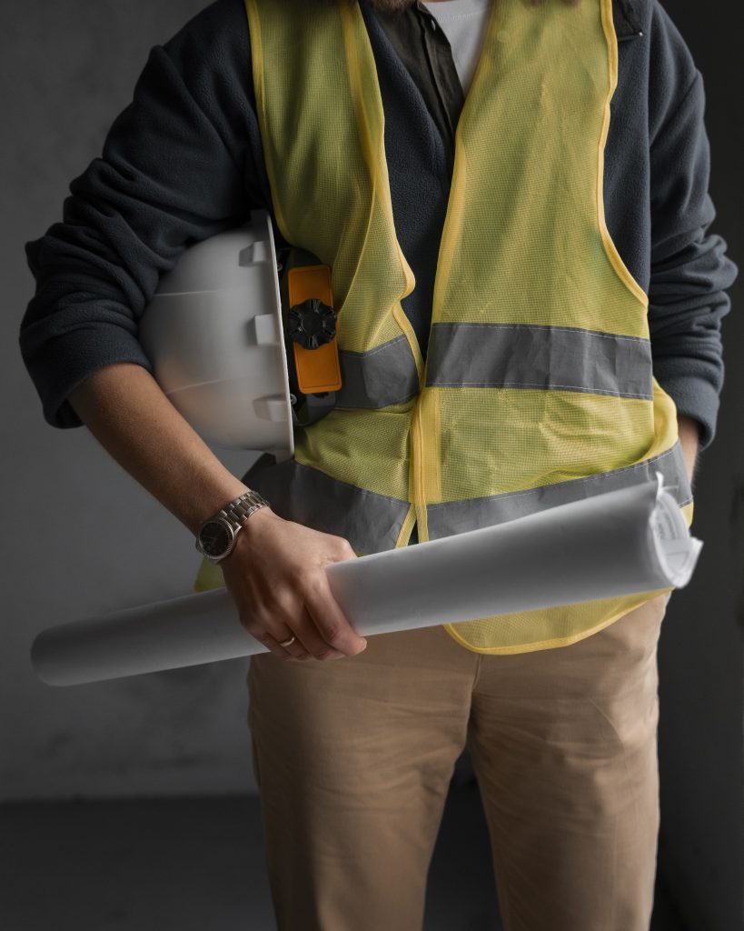A licensed contractor in Australia holding construction blueprints and a safety helmet, wearing a yellow safety vest and standing indoors.