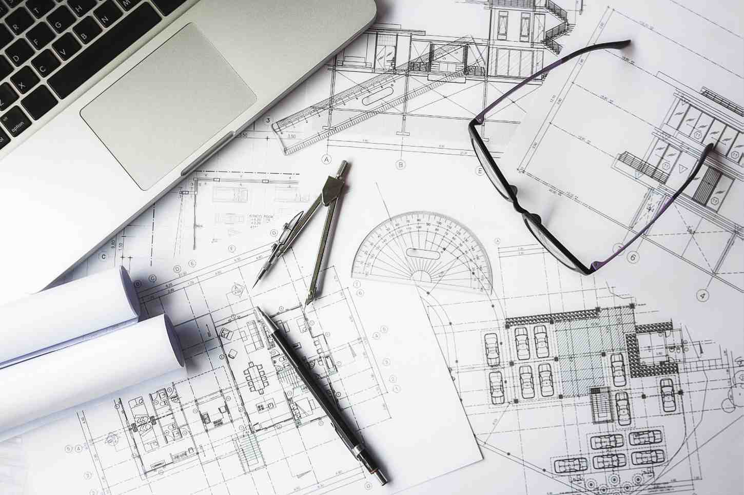 Construction blueprints on a desk with a laptop, compass, ruler, pen, and glasses, representing an engineer's workspace.