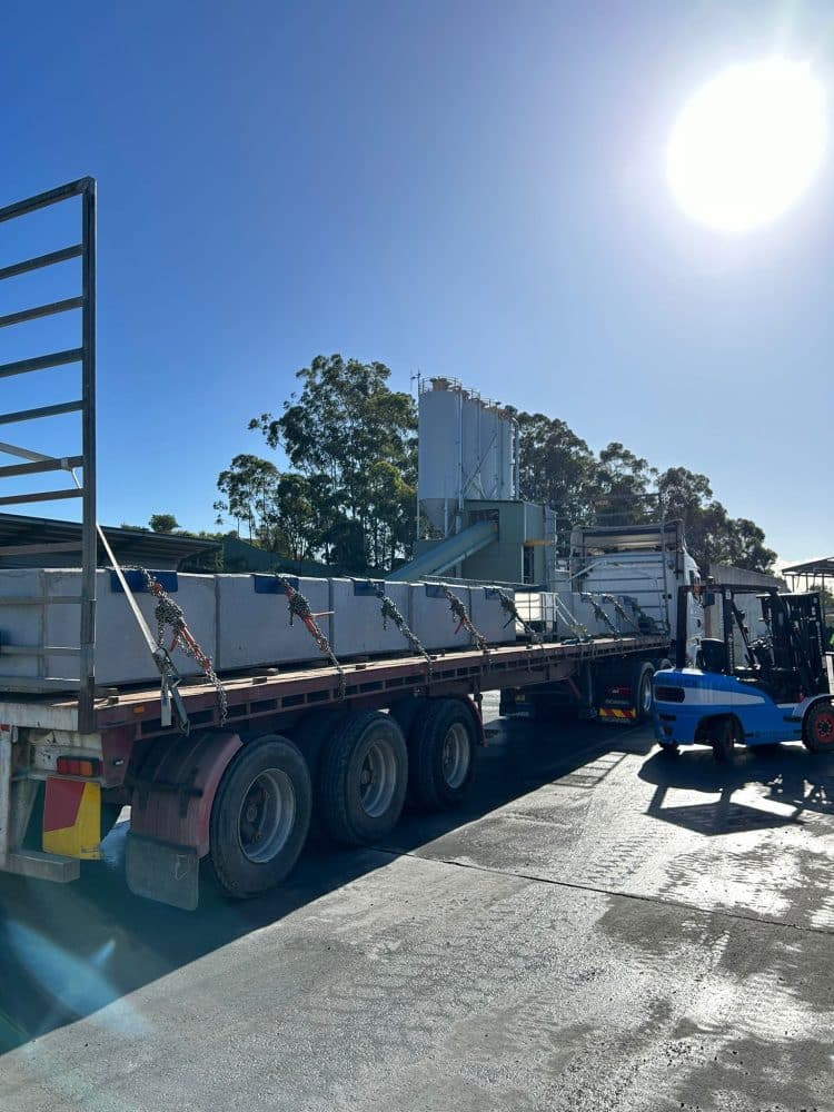 Flatbed truck loaded with precast concrete blocks at a site preparation area under bright sunlight.