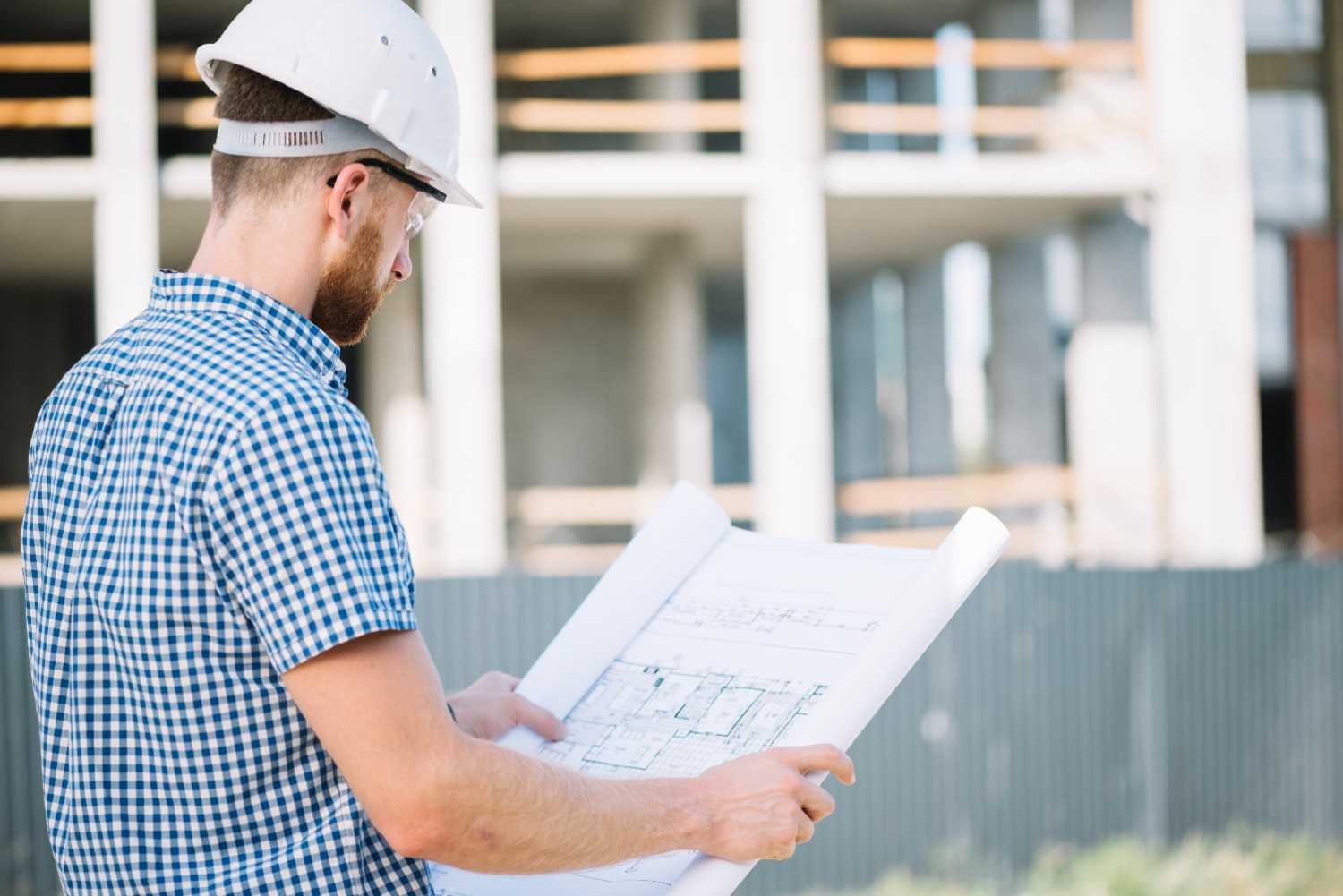 An architect wearing a hard hat examines a construction blueprint at a building site, with high-rise buildings in the background.