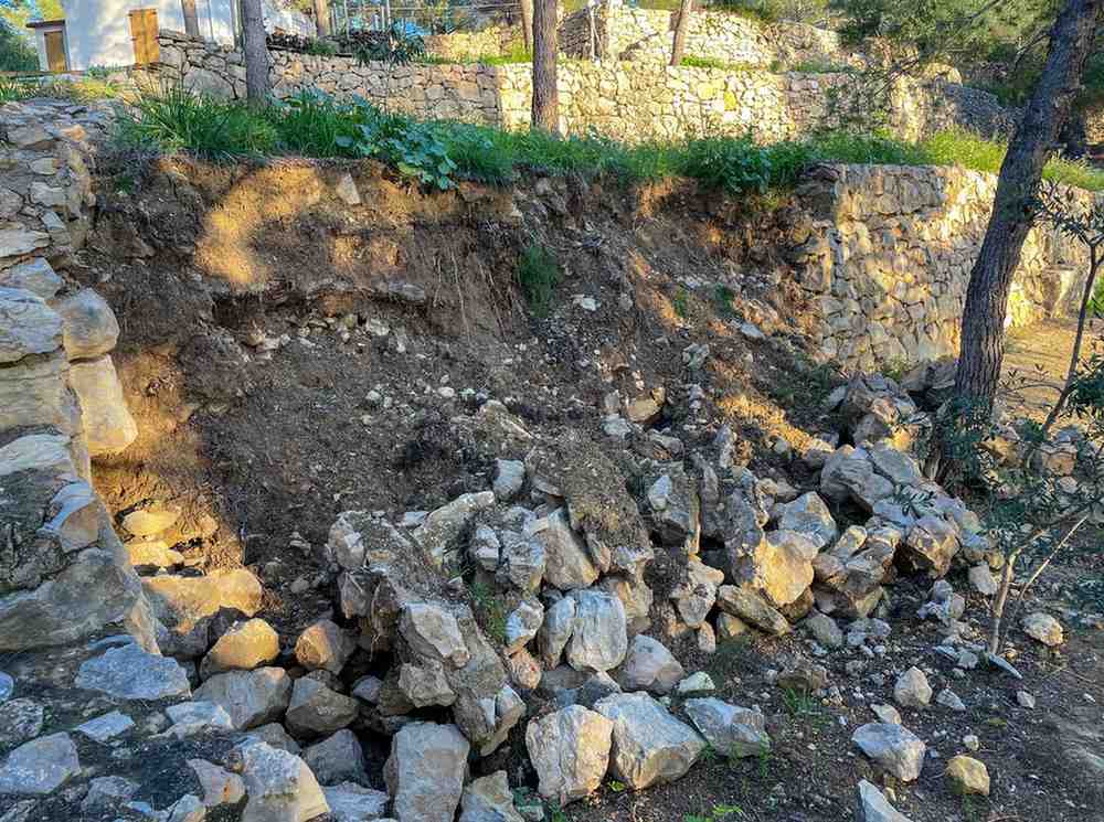 A collapsed dry-stone retaining wall with fallen rocks and exposed, eroded soil on a terraced slope
