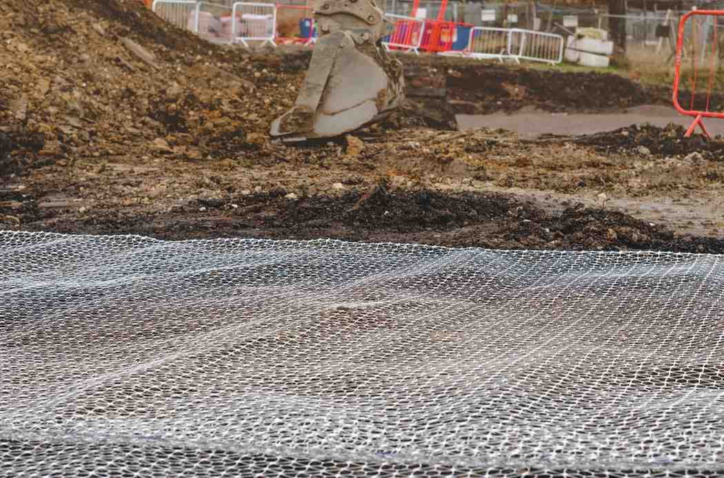 Geotextile separation mesh laid over excavated soil at a construction site, with an excavator and safety barriers behind