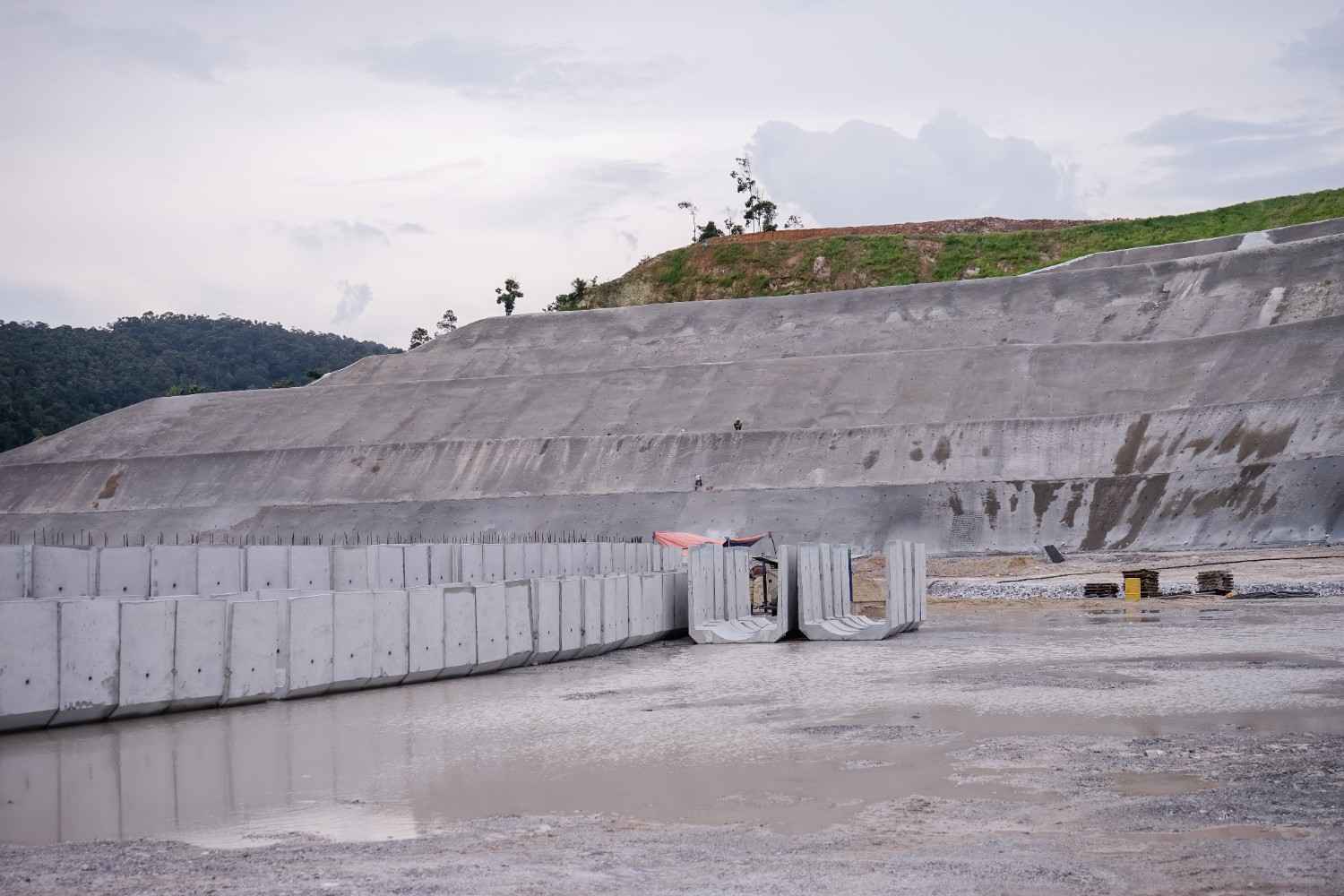 Precast concrete retaining wall panels set along a large civil earthworks site below a concrete-faced embankment