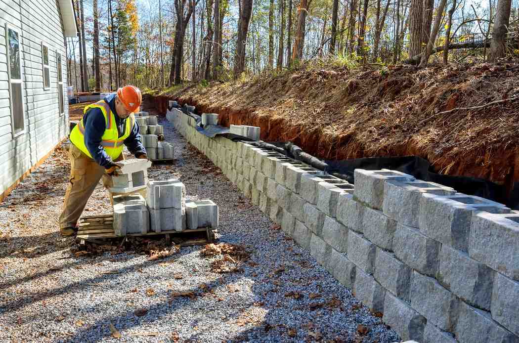 A worker building a concrete block retaining wall against a cut slope, with drainage fabric lining the exposed soil bank behind it