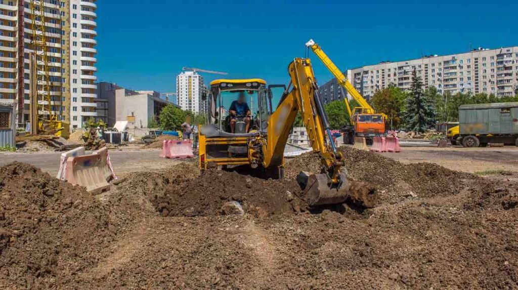 A backhoe loader excavating and turning over soil to prepare a construction site, with apartment buildings behind under a clear sky