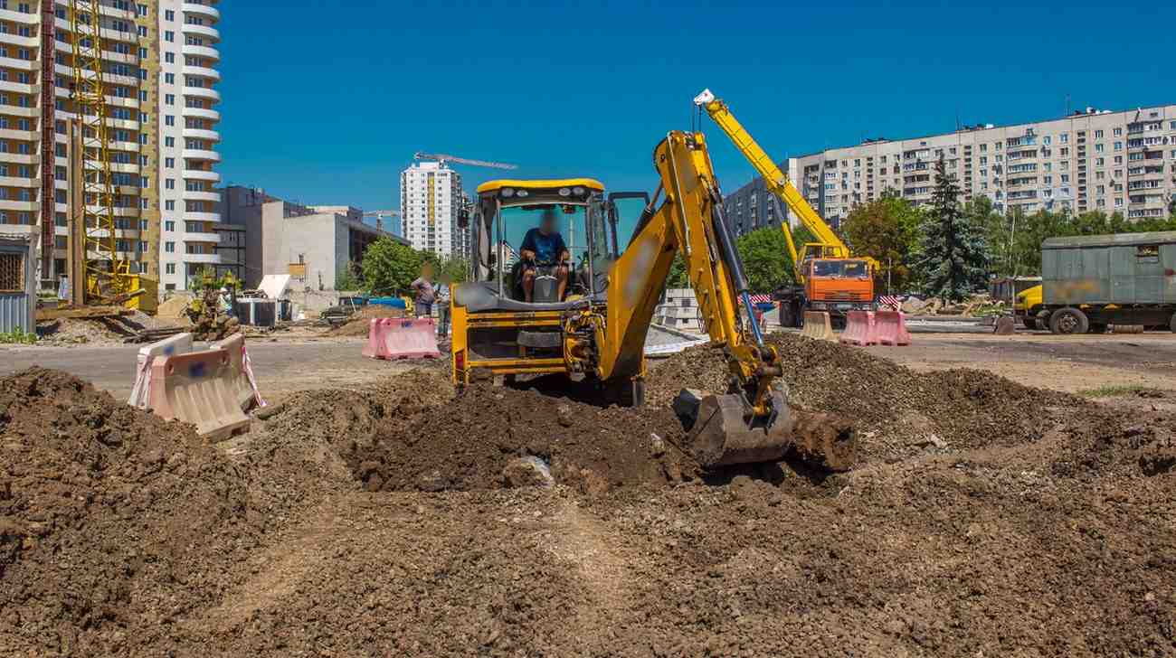 A backhoe loader excavating and turning over soil to prepare a construction site, with apartment buildings behind under a clear sky