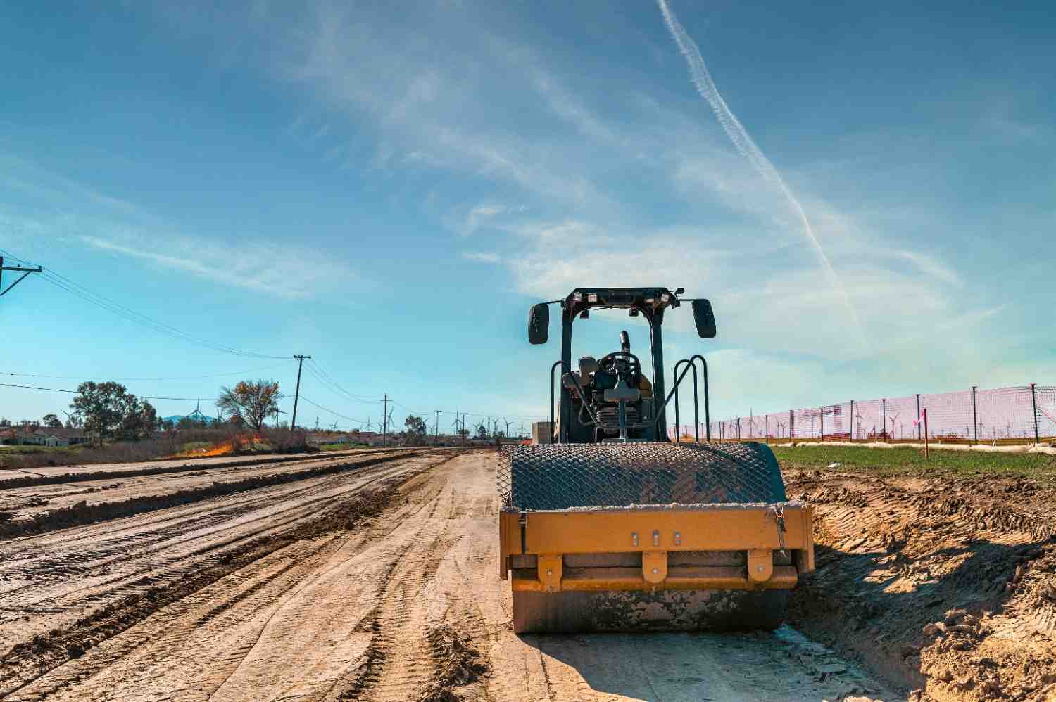 A single-drum roller compacting graded soil on a construction site under a clear sky