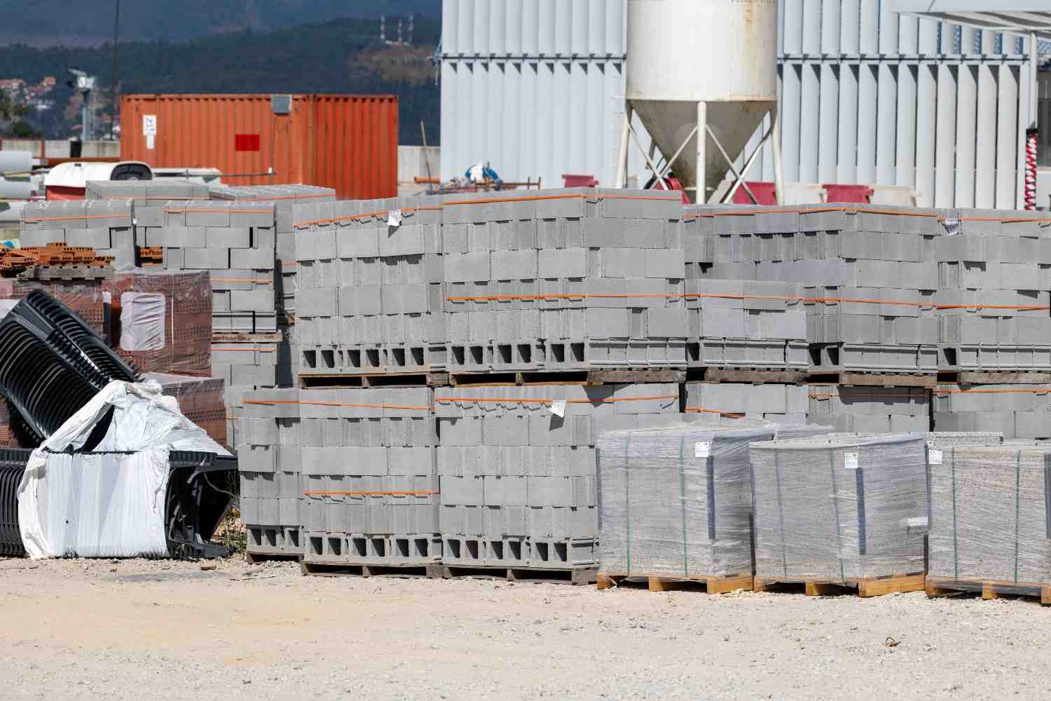 Pallets of grey concrete blocks stacked in an outdoor building-supply yard with cement silos behind